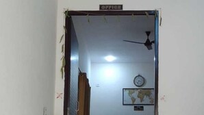 A hallway leading to an office area with a green carpet, a potted plant, a clock, and a ceiling fan. - Hotel CityMax Inn (Greater Noida)