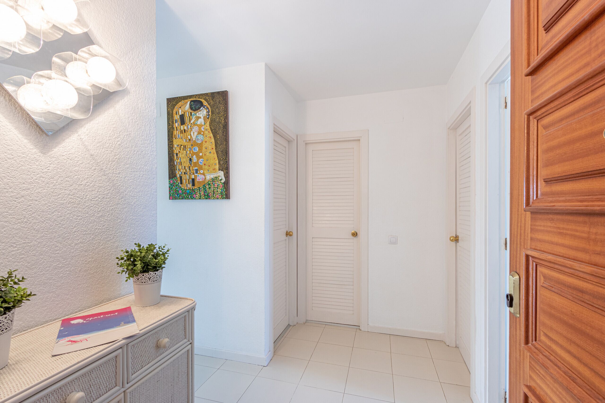 A hallway with white walls, a white tiled floor, a white dresser with two potted plants, a framed artwork, and closed doors.