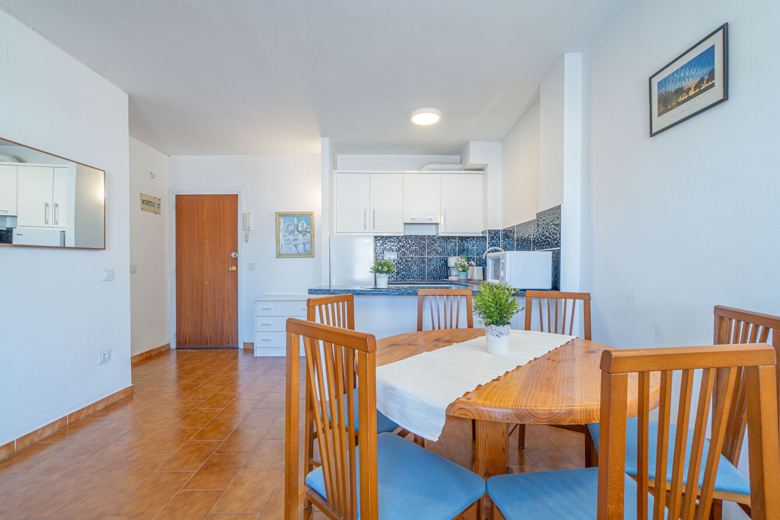 Een moderne keuken met witte kastjes, een betegelde spatwand en een houten eettafel met blauwe stoelen.