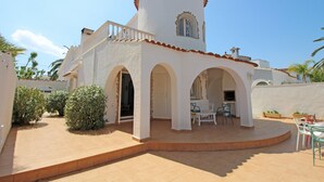 Un bâtiment blanc doté d’une tour ronde, de portes en arc de cercle et d’un patio aménagé avec des chaises et des tables.