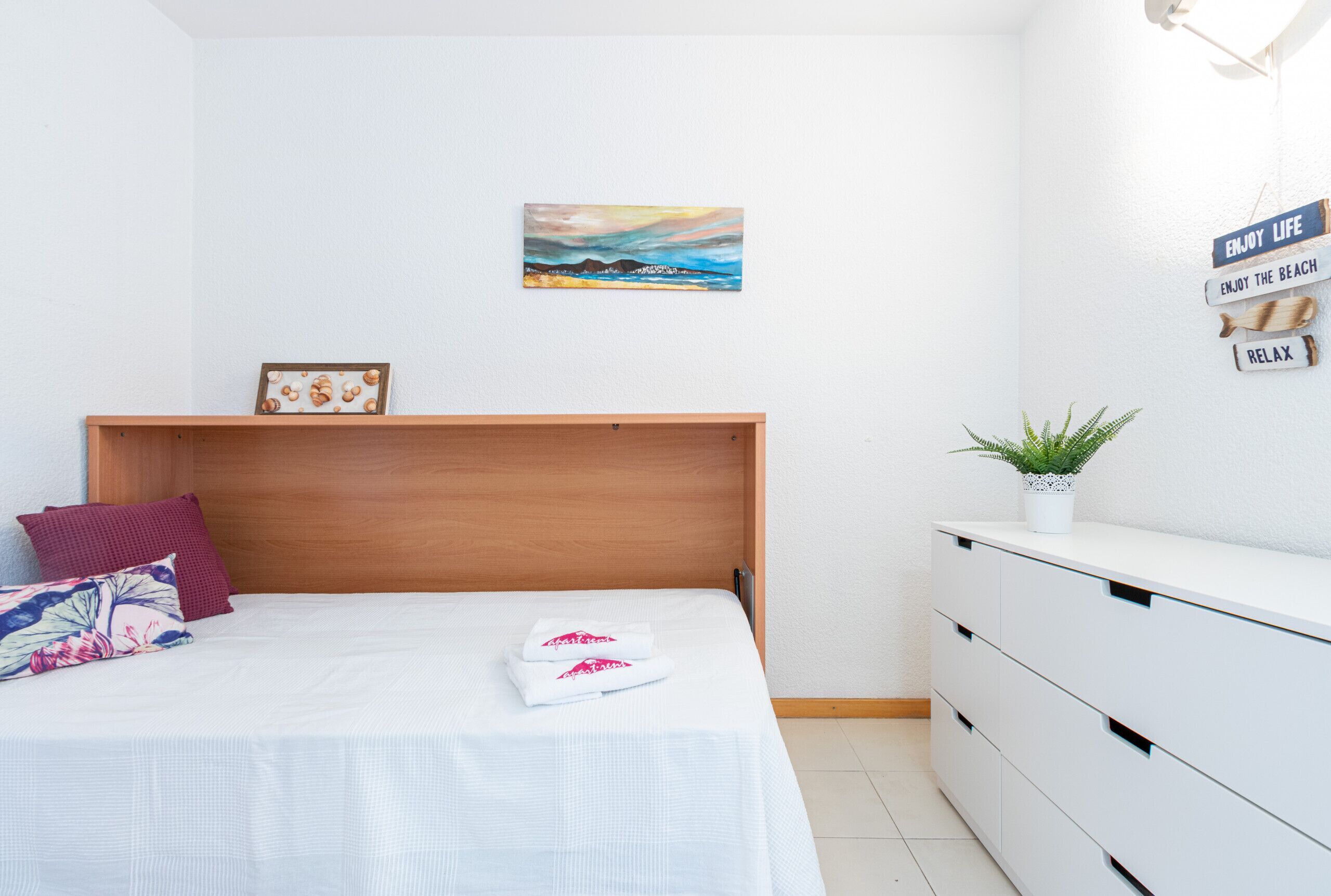 A bedroom with a wooden headboard, a bed with white bedding, a small framed picture, a potted plant, and a wall with motivational signs.