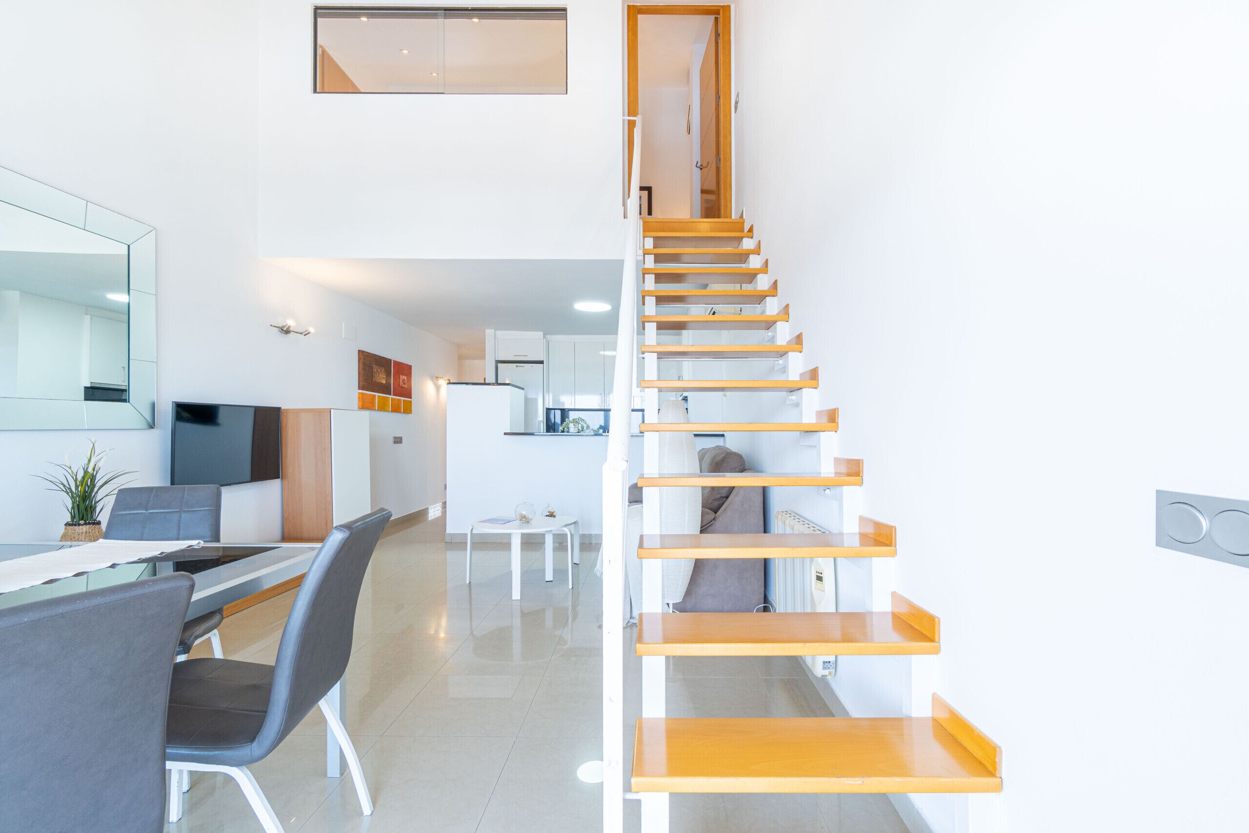 A modern kitchen with white cabinets, a dining area with a glass table, and a wooden staircase.