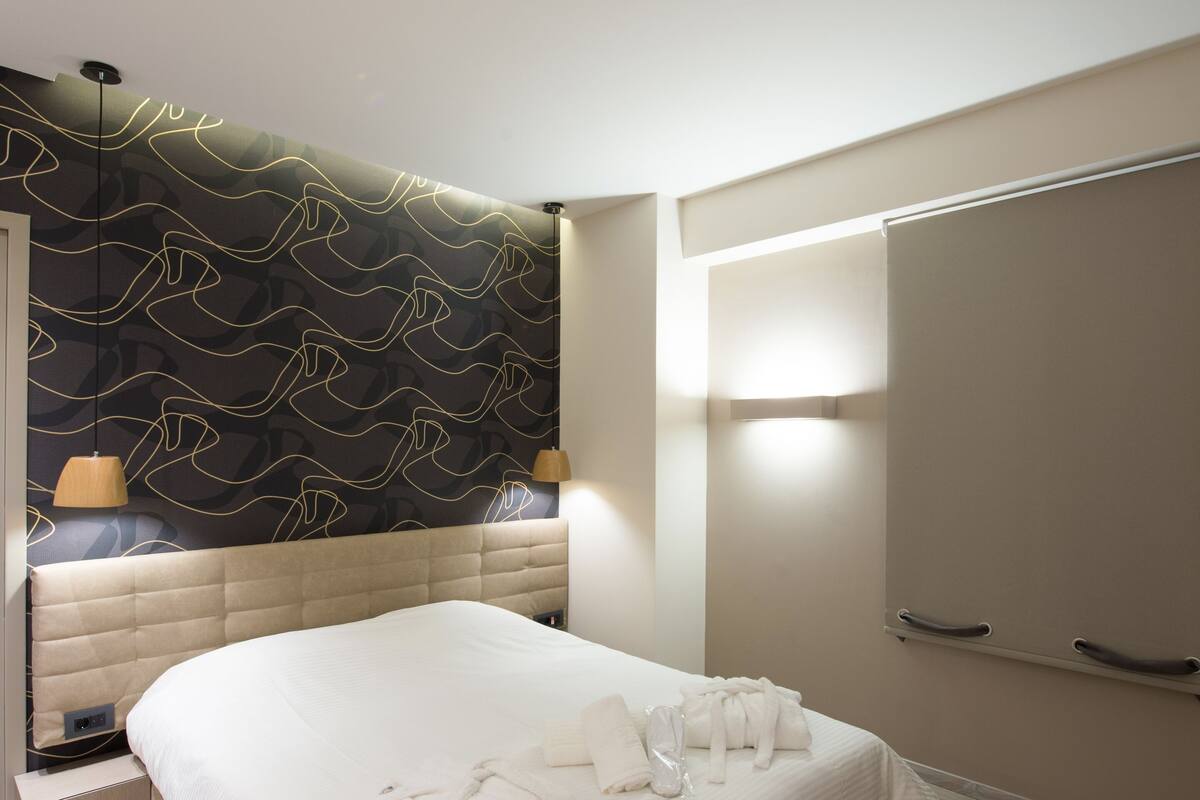 Quarto de hotel moderno com cama grande, cabeceira com padrão ondulado, mesa de cabeceira e guarda-roupa.