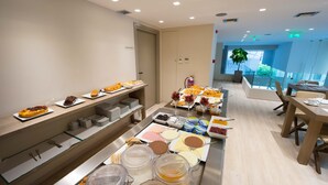 Un buffet con diversi cibi e bevande, un'area salotto con tavoli e sedie, e vista sul verde attraverso ampie finestre.