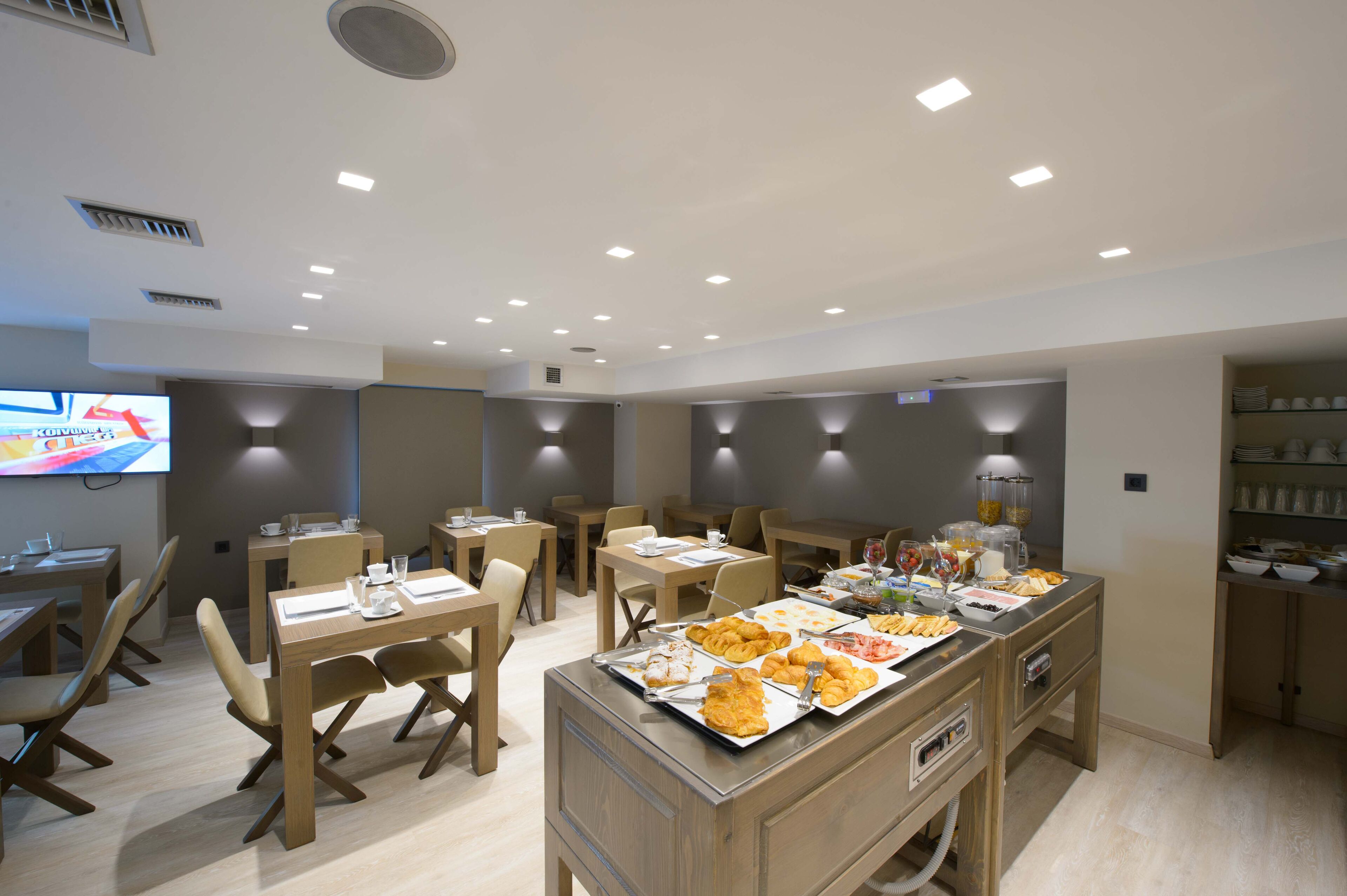 A modern dining area with wooden tables and chairs, a buffet with various food items, and a television screen on the wall.