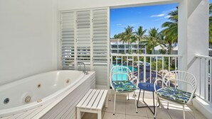 A balcony with a white bathtub, two chairs with tropical cushions, and a view of a pool and palm trees.