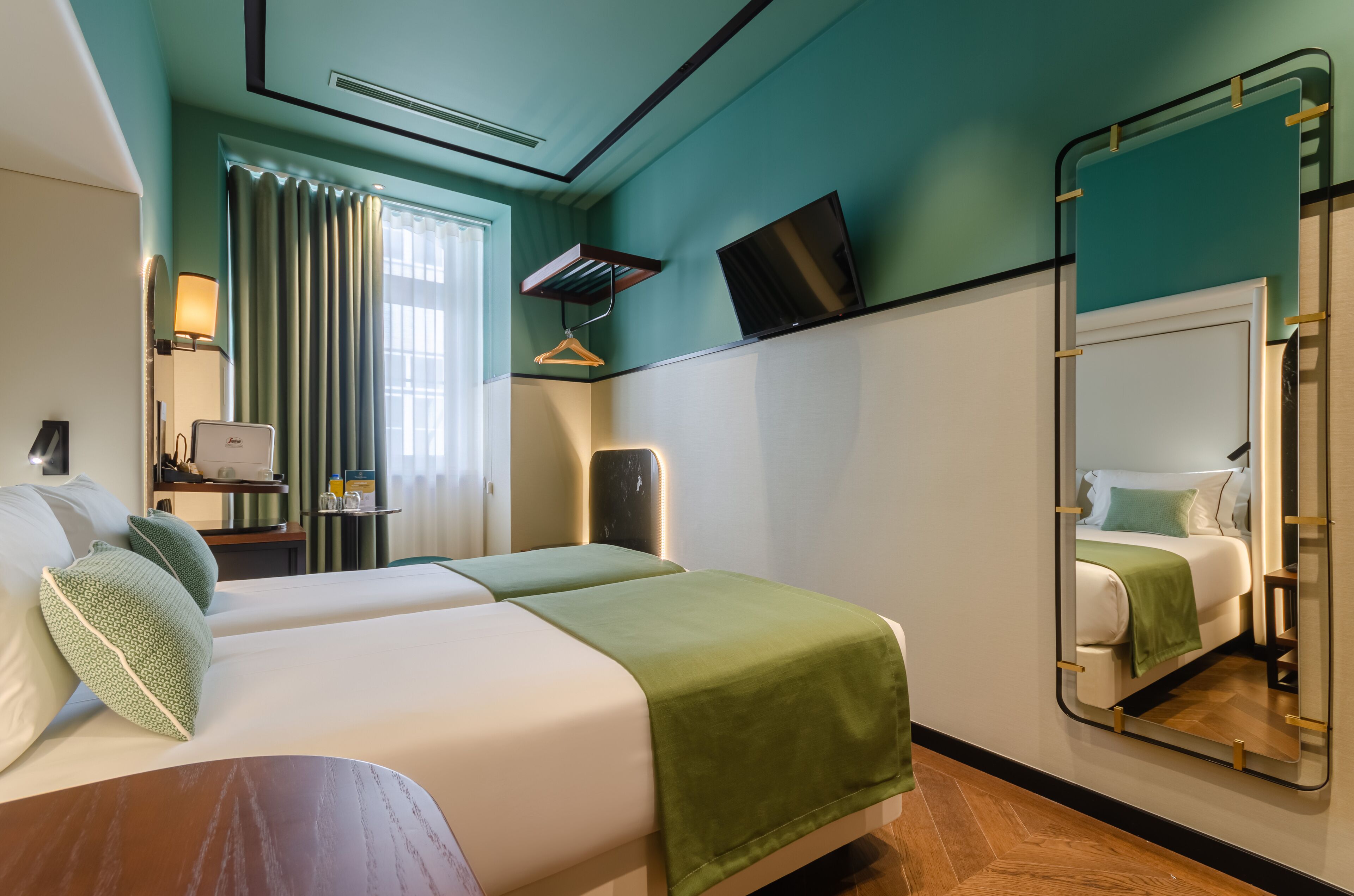 Quarto de hotel com duas camas, televisão, espelho, secretária com luminária e janela com cortinas.