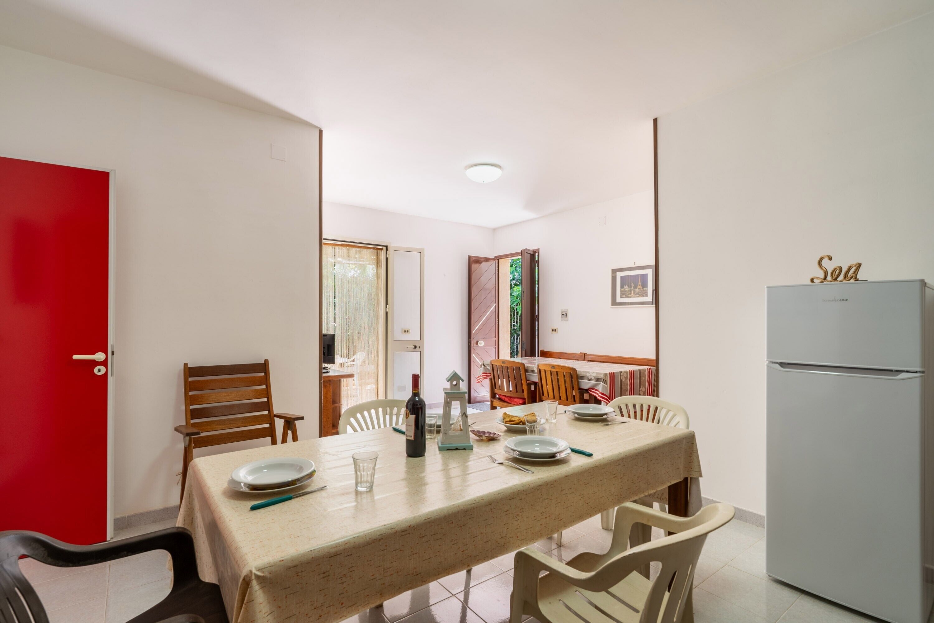 Un coin repas avec une table dressée pour deux personnes, des chaises, un réfrigérateur et une porte rouge.