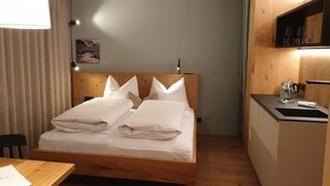 Un lit simple avec du linge de lit blanc, une tête de lit en bois, une table de chevet avec une lampe, un petit coin cuisine avec un évier et une bouilloire, et une fenêtre avec des rideaux.