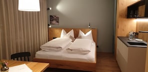 Un letto singolo con lenzuola bianche, una testiera in legno, un comodino con una lampada, un angolo cottura con lavello e bollitore, e una finestra con tende.