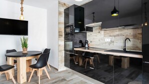 A modern kitchen with a dining area, featuring a wooden table, black chairs, a flat-screen TV, and a built-in oven. - Casa Bombón - Country House with Private Pool & Wi-Fi – Families Only (Vejer de la Frontera)