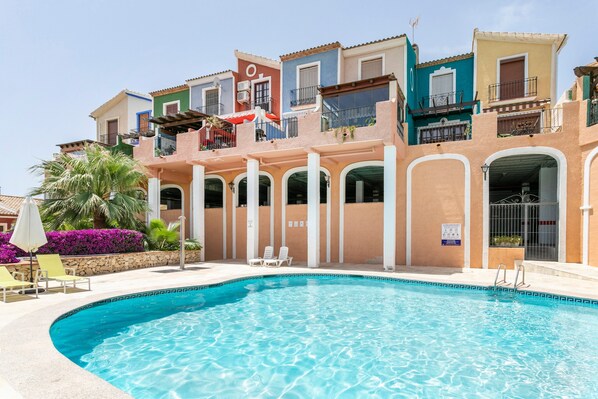 Une piscine avec des chaises longues, un palmier et des bâtiments aux couleurs vives en arrière-plan.