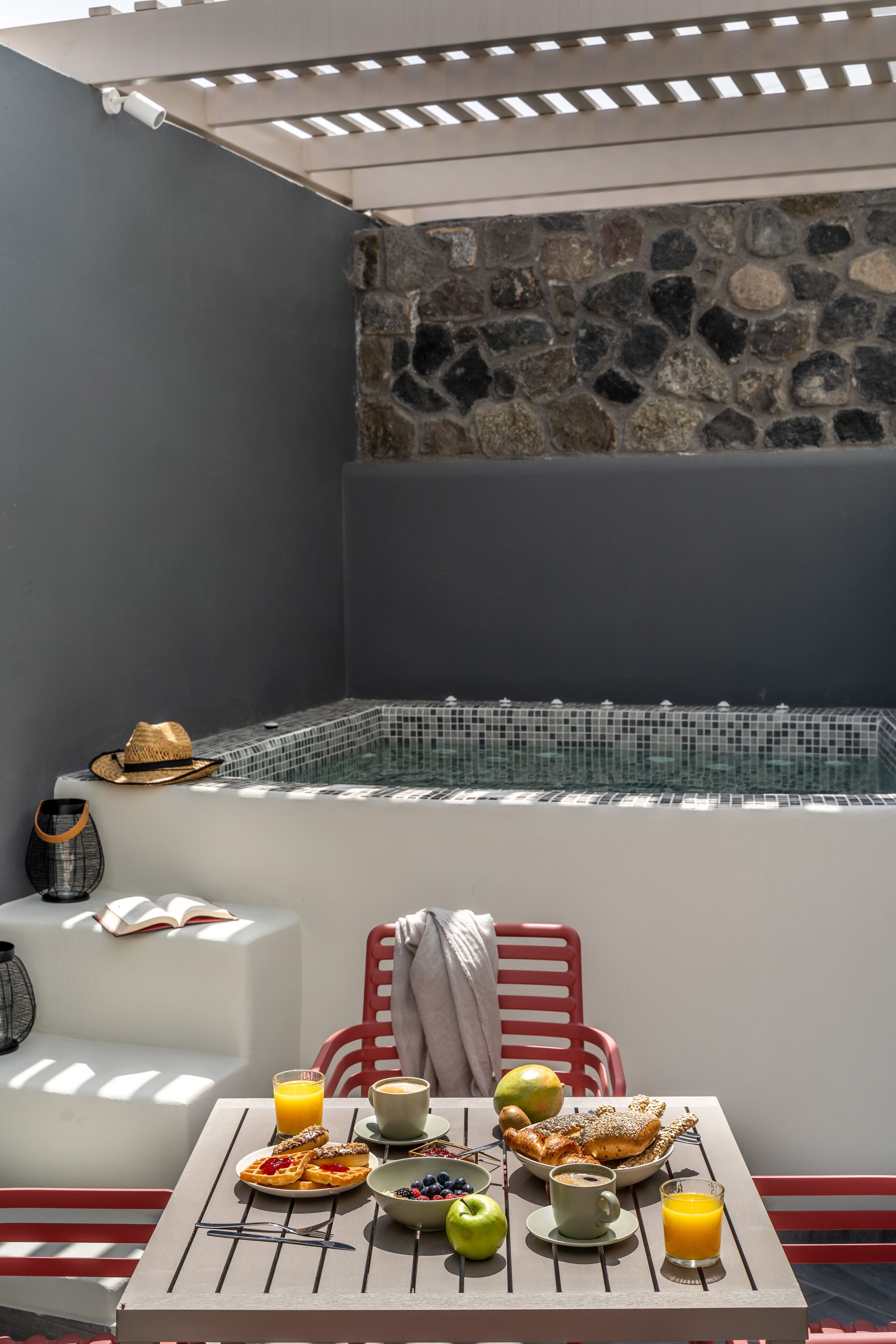 A cozy outdoor seating area with a stone wall, a white cushioned bench, and a table set with breakfast items including toast, fruit, and a drink.