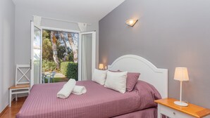 Een tweepersoonsbed met een roze sprei, een nachtkastje met een lamp, een stoel, en uitzicht op groen via een geopende deur.