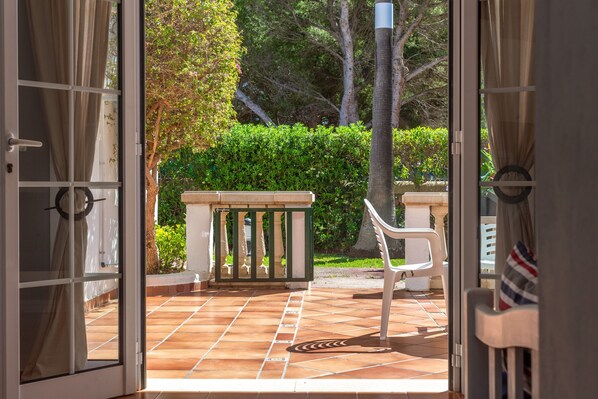 Une porte ouverte donne sur un patio où se trouvent une chaise blanche, une petite table et, au fond, une haie verte.