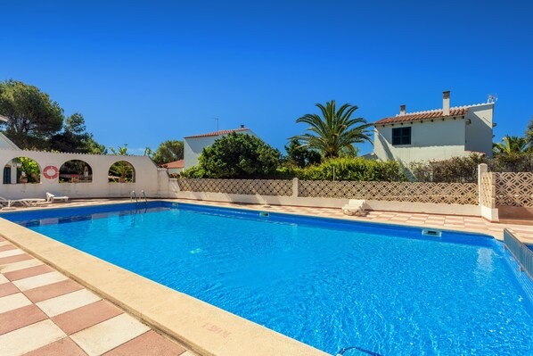 Une piscine à l’eau d’un bleu cristallin, entourée d’une terrasse carrelée et bordée d’un bâtiment blanc orné d’arcades.