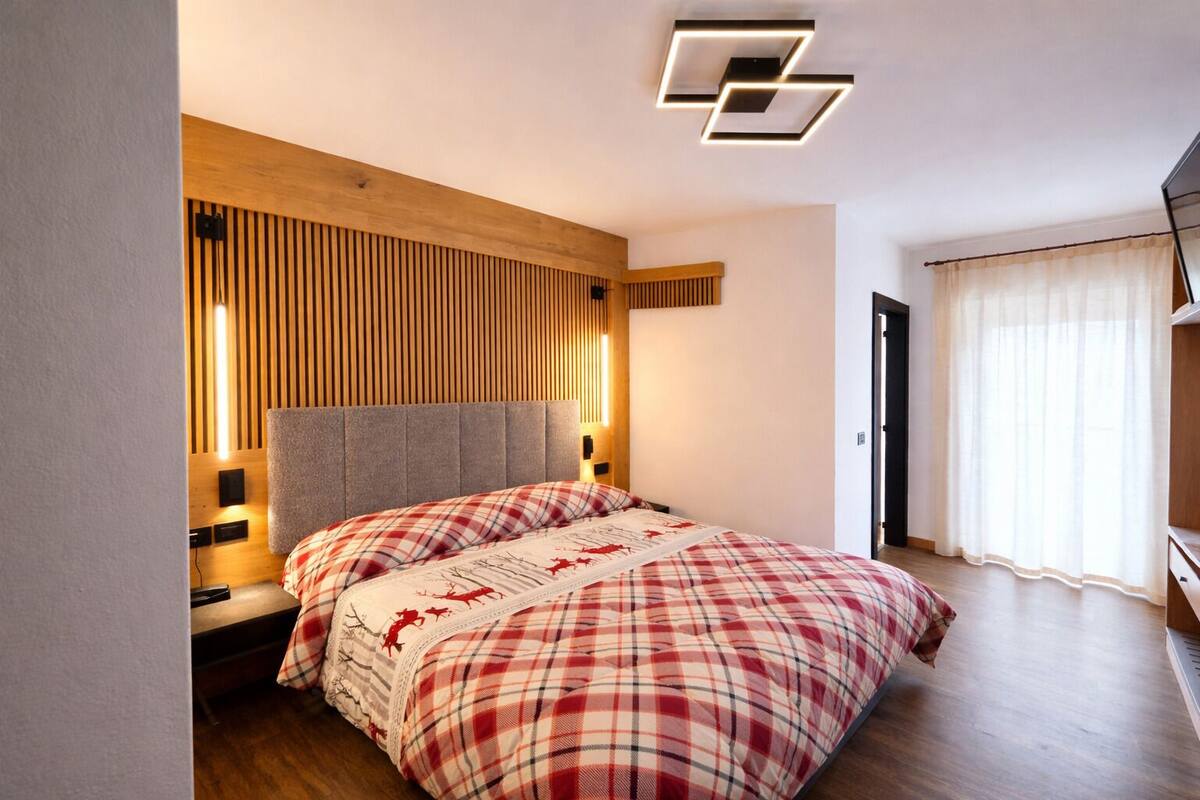 Une chambre à coucher avec un lit, un mur recouvert de panneaux en bois, un luminaire moderne au plafond et une fenêtre avec des rideaux.