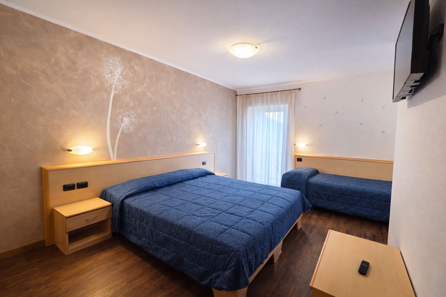 Habitación de hotel con dos camas, una mesita de noche de madera, una pequeña mesa y un televisor montado en la pared.