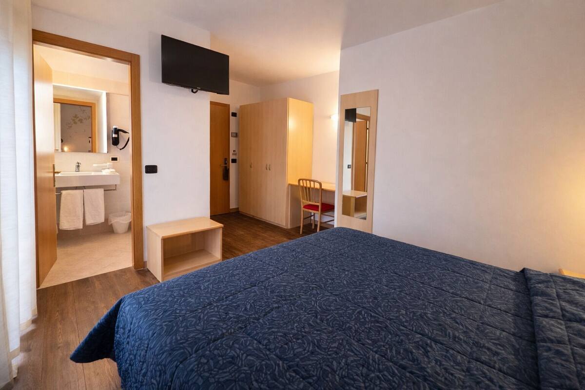 Une chambre à coucher comprenant un lit, un bureau, une chaise, un téléviseur et une salle de bain visible à travers une porte ouverte.