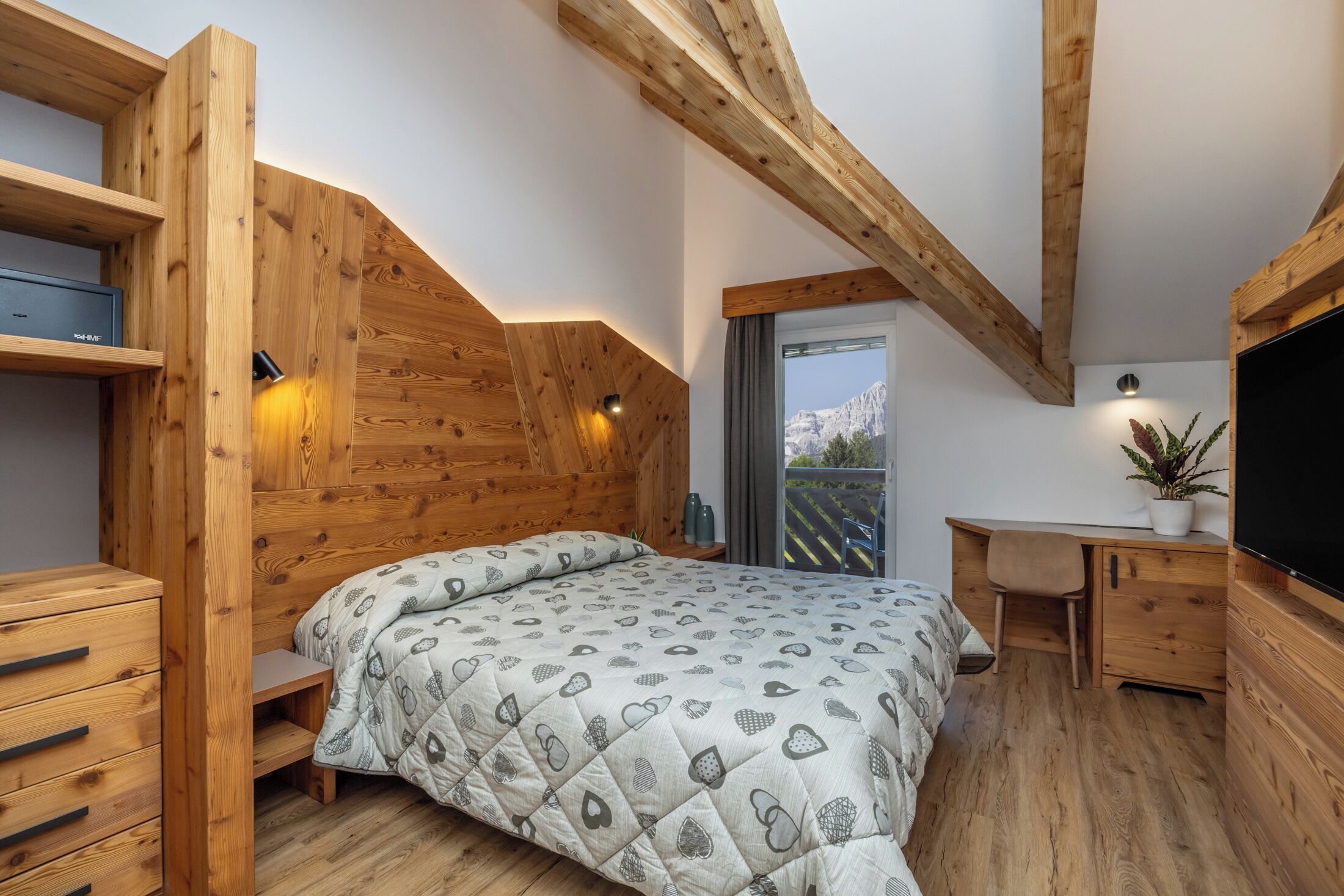 A bedroom with a wooden sloped ceiling, a bed with a patterned duvet, a desk, a chair, and a TV.