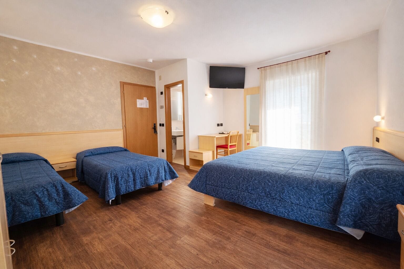 Ein Hotelzimmer mit drei Betten, einem Schreibtisch, einem Stuhl, einem Fernseher und einem Badezimmer.