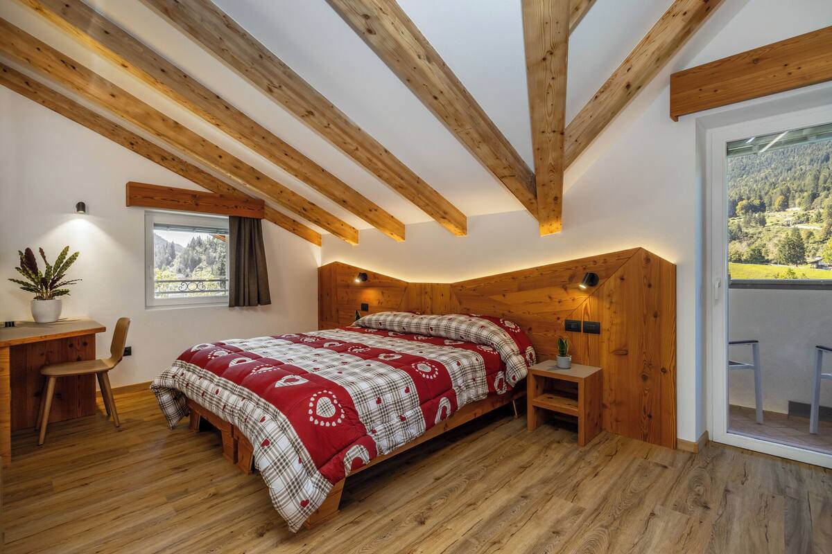 Une chambre avec un plafond en bois, un lit recouvert d’une couette à carreaux rouges et blancs, un bureau avec une chaise et une fenêtre donnant sur une forêt.