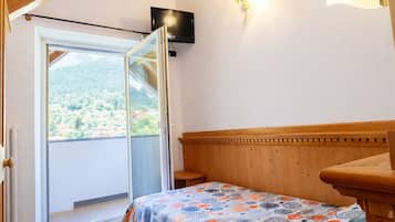 Ein Schlafzimmer mit Holzbalkendecke, einem Bett mit gemustertem Überwurf, einem Nachttisch, einem an der Wand montierten Fernseher und einem Fenster mit Blick auf eine Berglandschaft.
