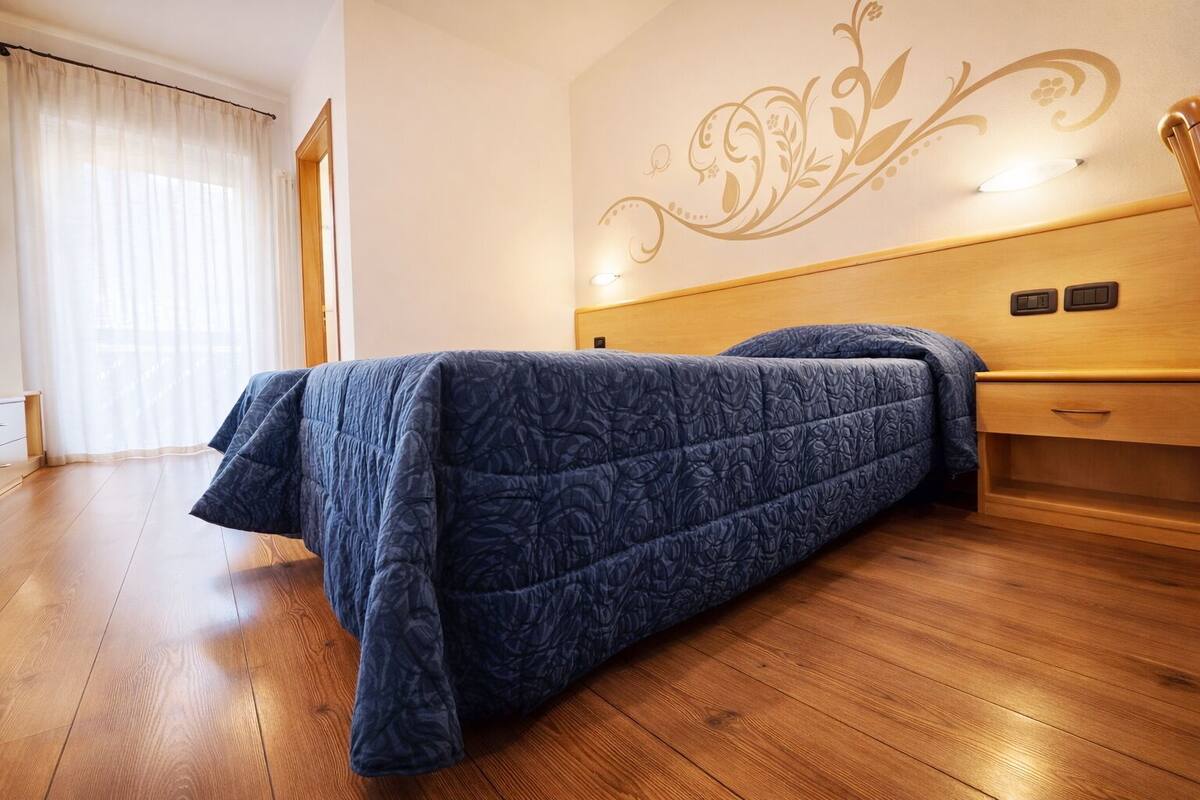 Une pièce avec un lit, un plancher en bois, un mur orné d’un motif floral et une fenêtre avec des rideaux.