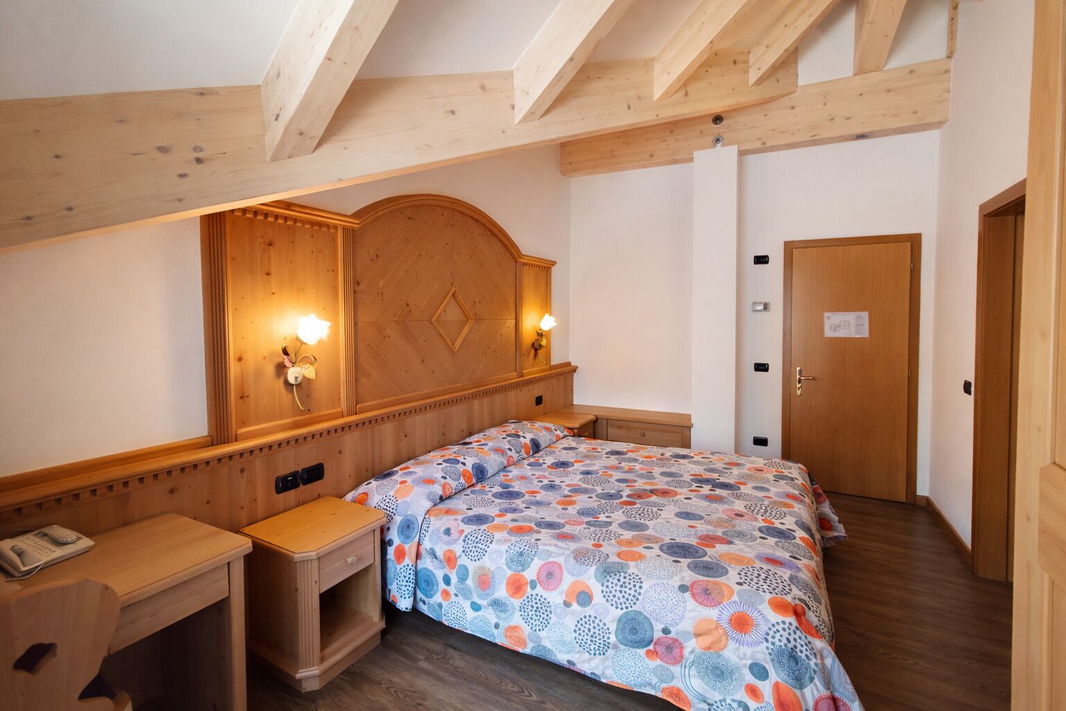 Ein Hotelzimmer mit einem hölzernen Kopfende, einem Bett mit einem gemusterten Bettbezug, einem Nachttisch mit Telefon und einer Holztür.