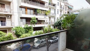 A balcony with a view of a street, parked cars, and multi-story buildings with balconies and plants.