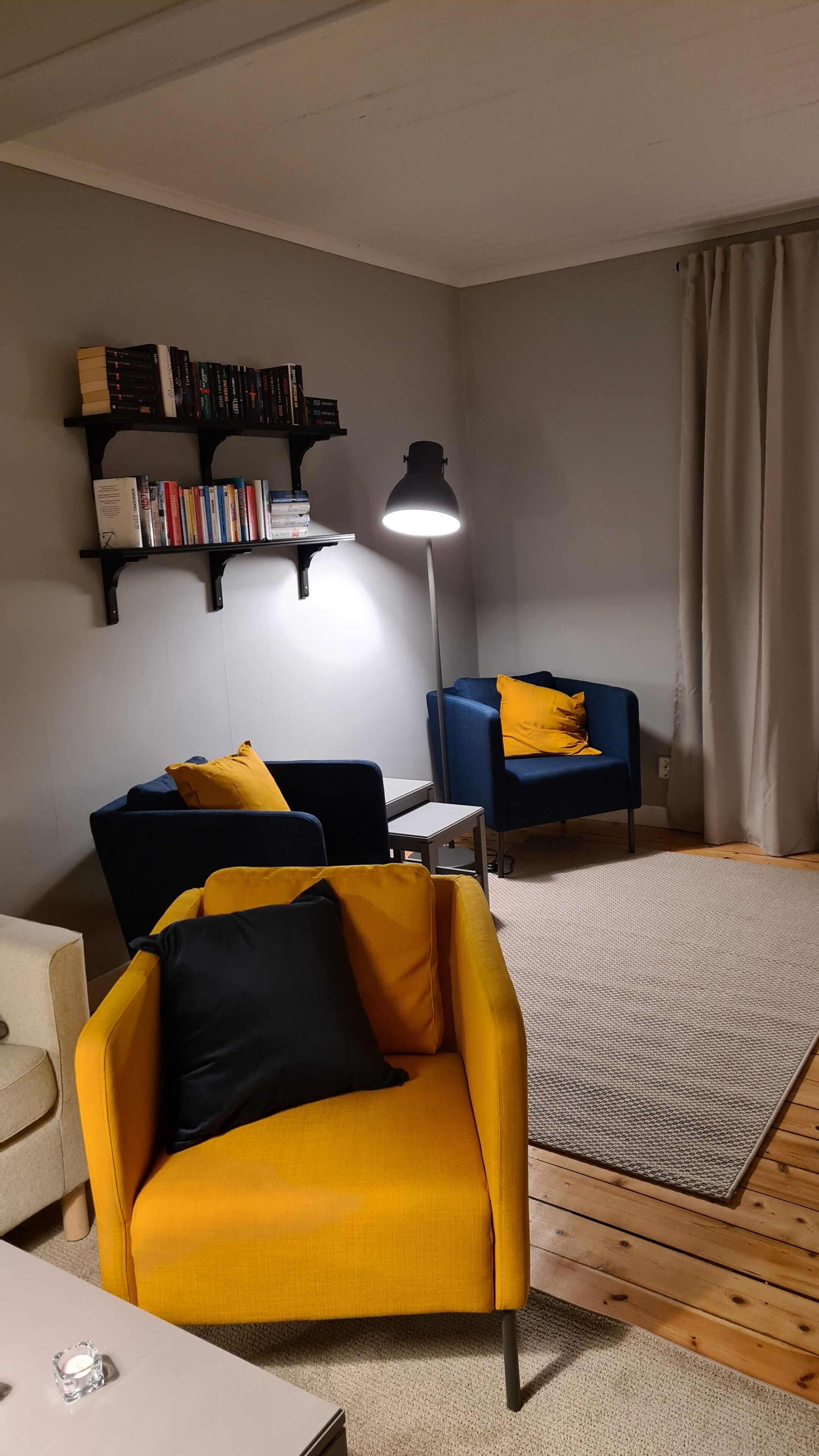 A living room with a yellow armchair, a blue sofa, a floor lamp, and a bookshelf.