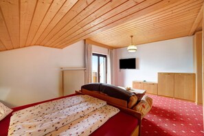 Un dormitorio con techo de madera, una cama con colcha roja estampada con motivos florales, un armario de madera y una ventana con cortinas.