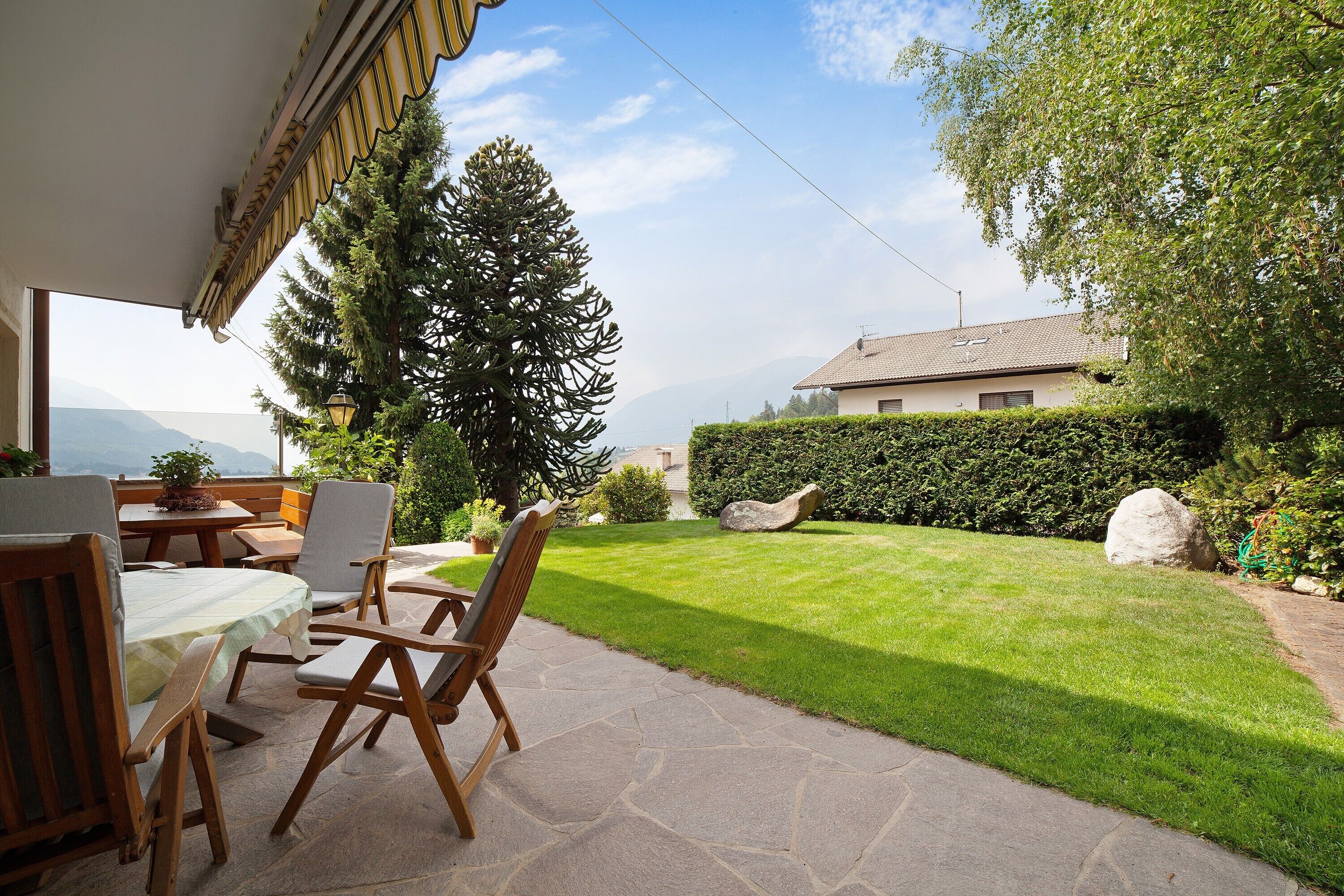 Eine Terrasse im Freien mit Steinböden, Holzmöbeln und Blick auf einen gepflegten Rasen und ferne Berge.