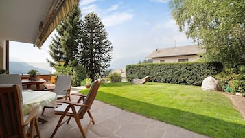 Eine Terrasse im Freien mit Steinböden, Holzmöbeln und Blick auf einen gepflegten Rasen und ferne Berge.