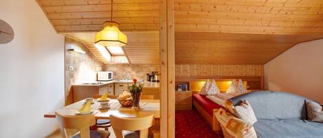 Una habitación acogedora con techo de madera, un comedor con sillas y mesa, un sofá con cojines, y una cocina al fondo.