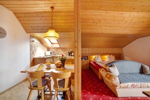 A cozy room with a wooden ceiling, a dining area with chairs and a table, a sofa with cushions, and a kitchen in the background.
