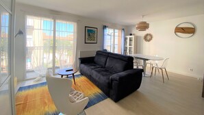 A spacious living room with a black sofa, a small round table, and a dining area with a white table and chairs. - Central Arcachon - Bright 3* Duplex T3 with Balcony, Parking, and Modern Amenities (Arcachon)
