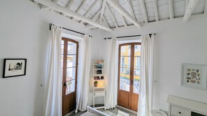 Een kamer met witte wanden, een houten plafond en twee ramen met houten kaders en witte gordijnen. Er staat een bed met een wit hoofdeinde, een klein witte dressoir en een plank met boeken en decoratieve objecten.