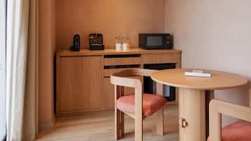 A compact kitchenette with a microwave, coffee maker, and cabinets. There is a small round table with a chair and a book on it. The room has wooden flooring and beige walls.