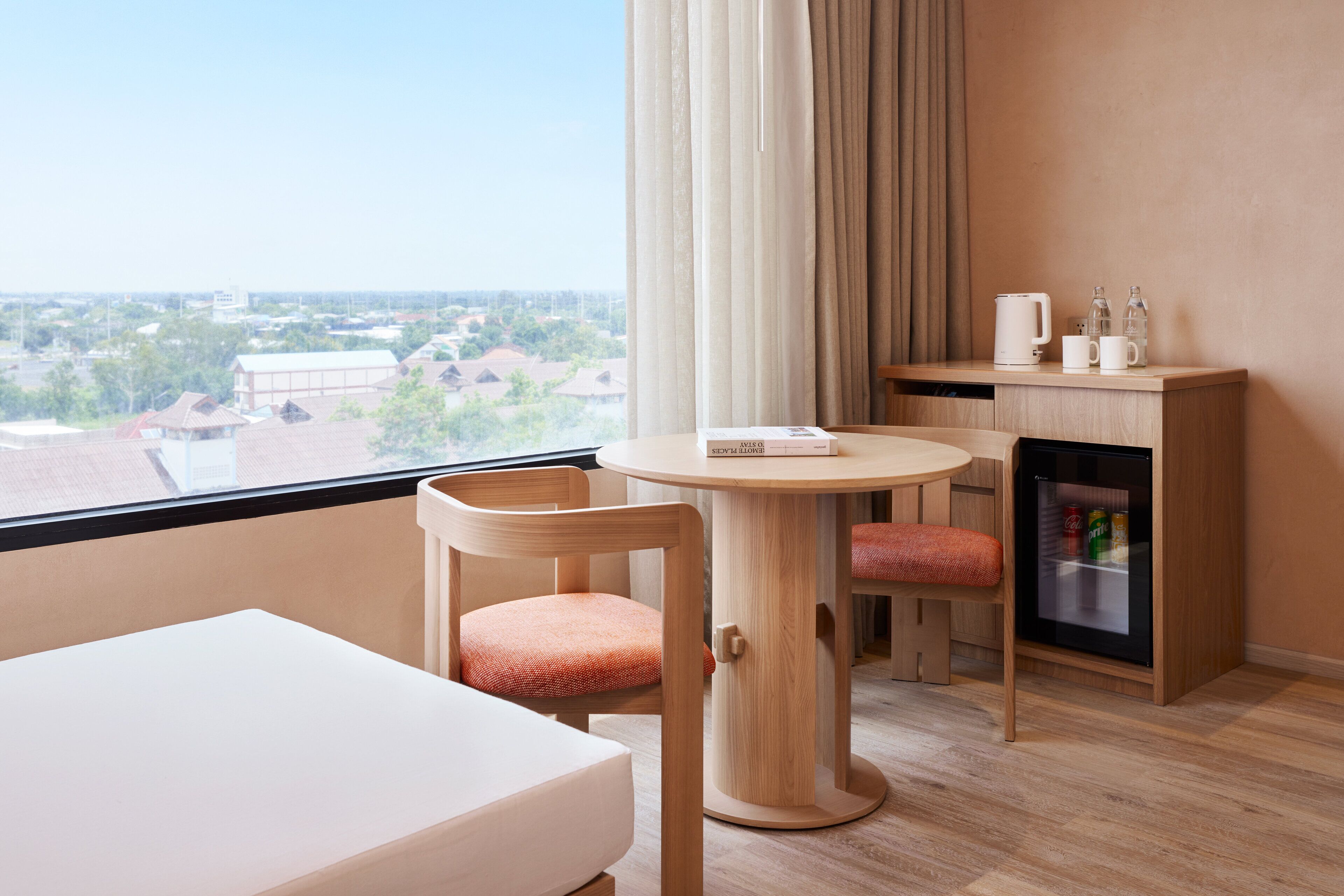 A modern hotel room with a large window, a round table, two chairs, a minibar, and a white bed.