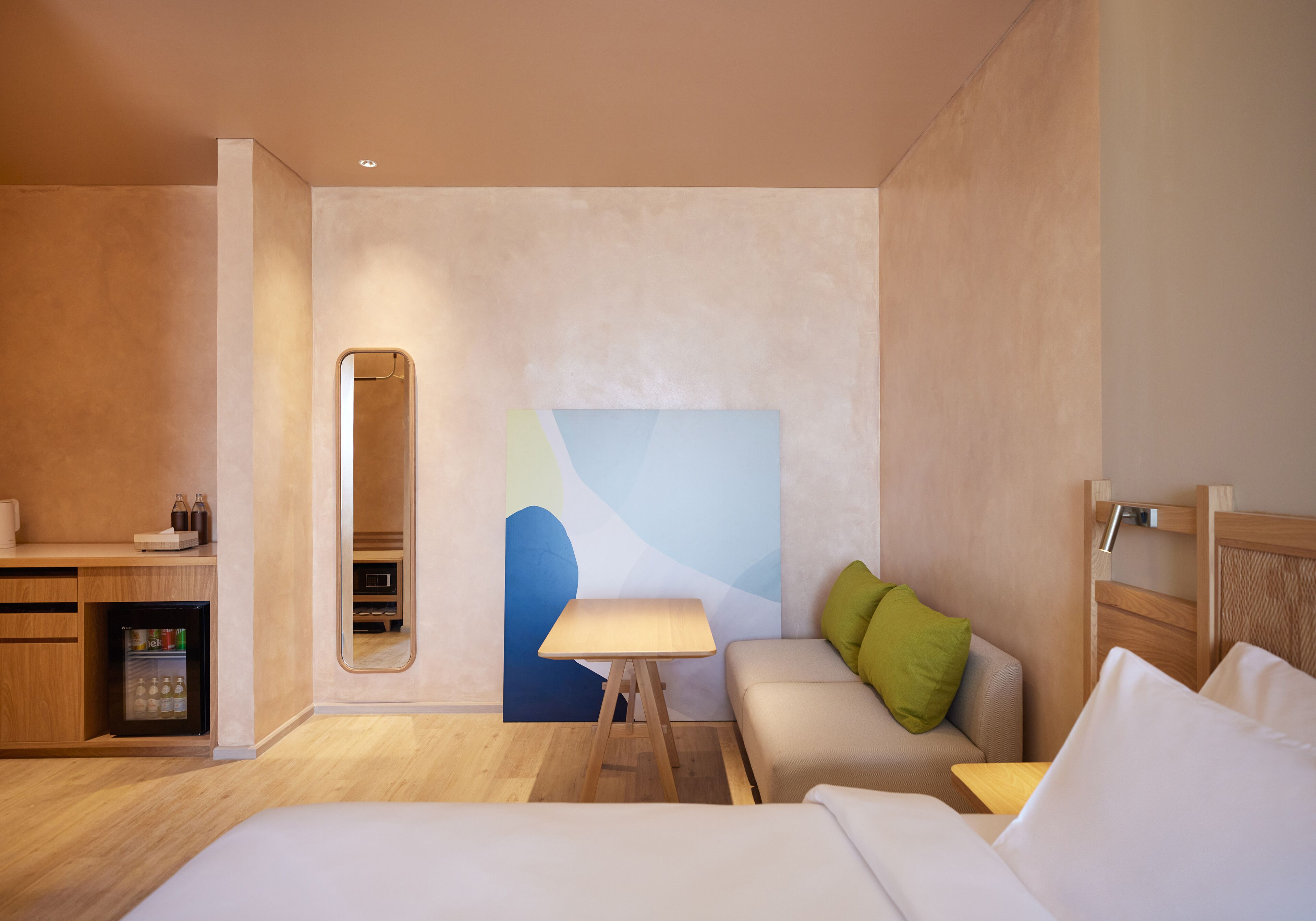 A modern hotel room with a bed, a small desk with a refrigerator, a seating area with a sofa and a table, and a large abstract painting on the wall.