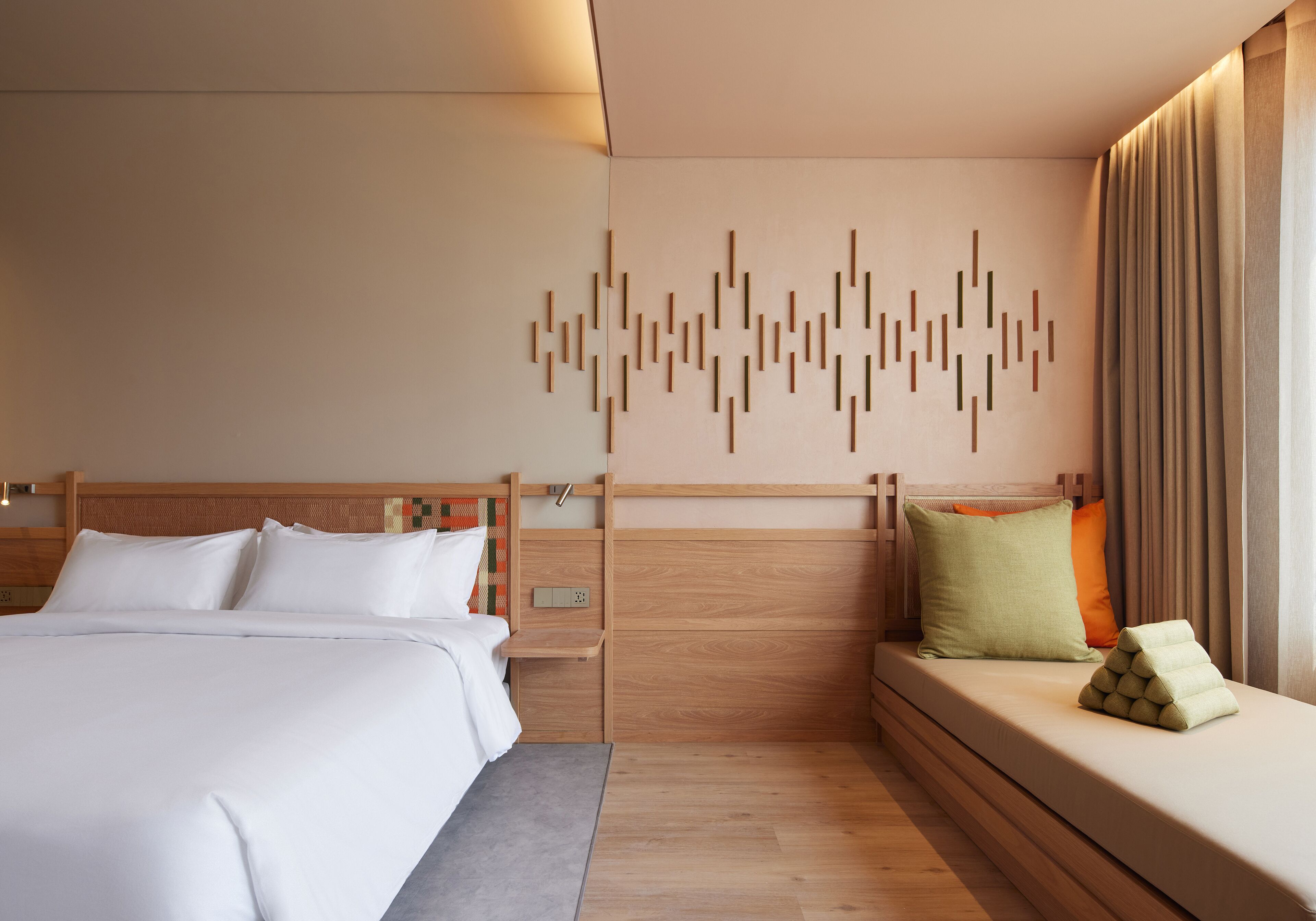 A modern hotel room with a wooden headboard, a bed with white linens, a bedside table, a built-in bench with cushions, and a wall-mounted light fixture.