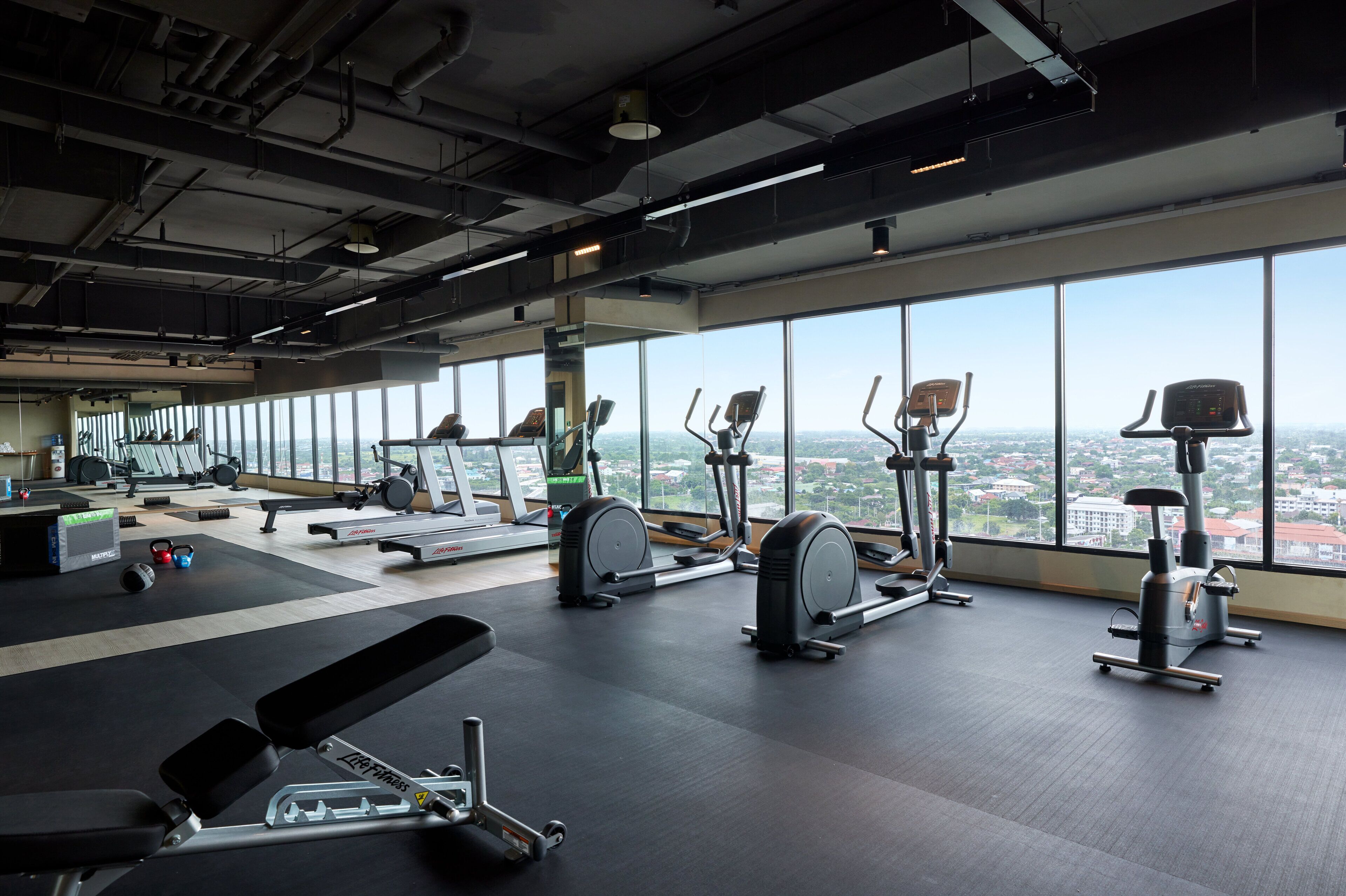 Un gimnasio con diverso equipo de ejercicio, amplias ventanas y vistas a la ciudad.