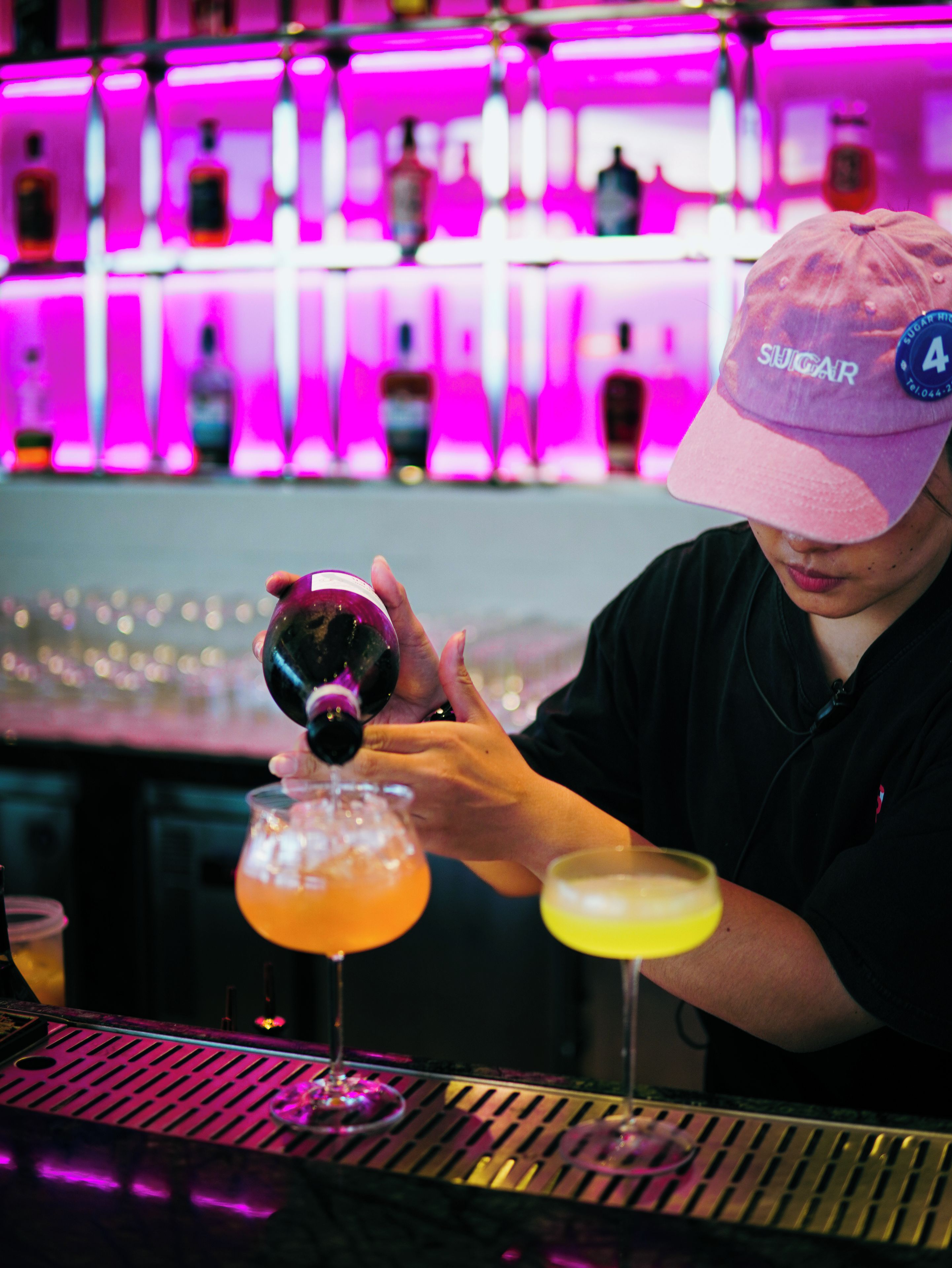 A person pouring a drink into a glass at a bar with a neon pink background.