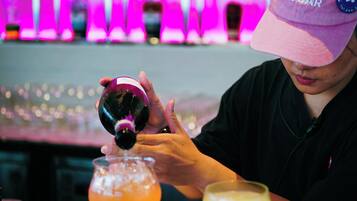 A person pouring a drink into a glass at a bar with a neon pink background.