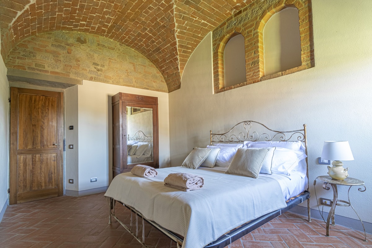 A bedroom with a brick ceiling, a bed with white linens, a wooden door, and a small table with a lamp.