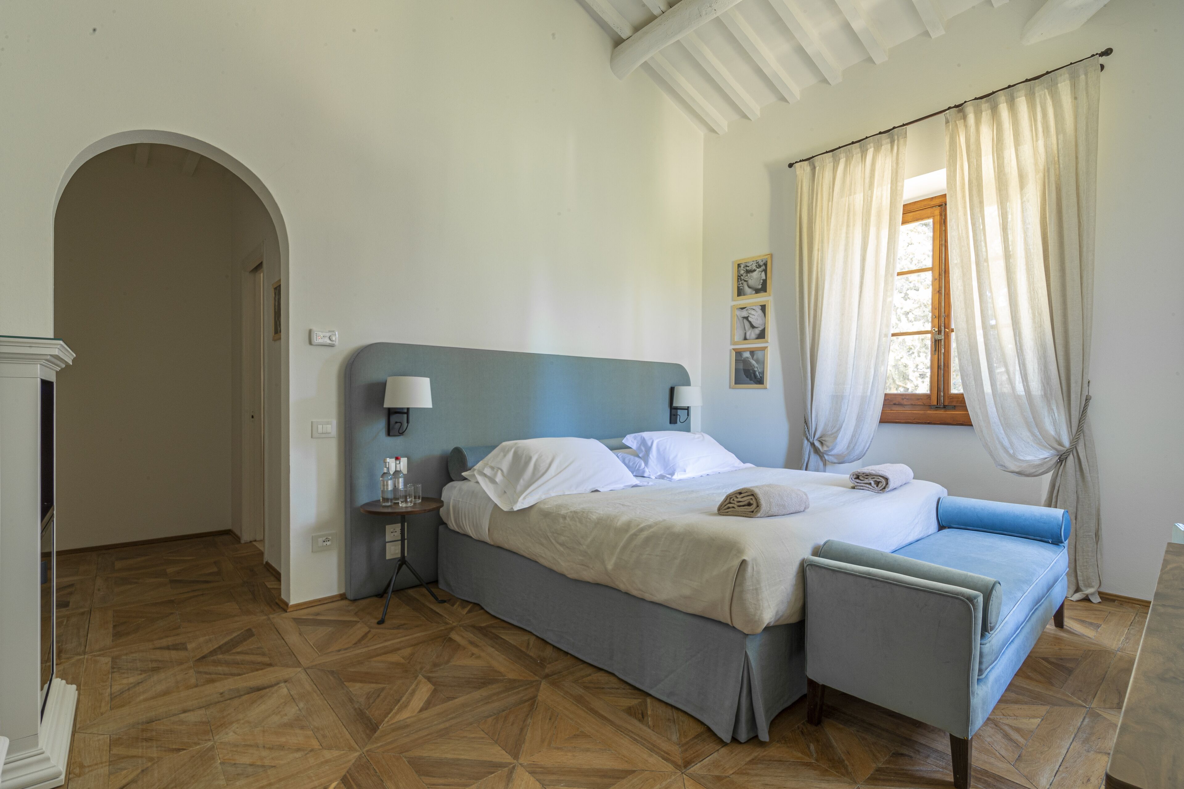 A bedroom with a bed, bedside tables, a chair, a window with curtains, and a wooden floor.