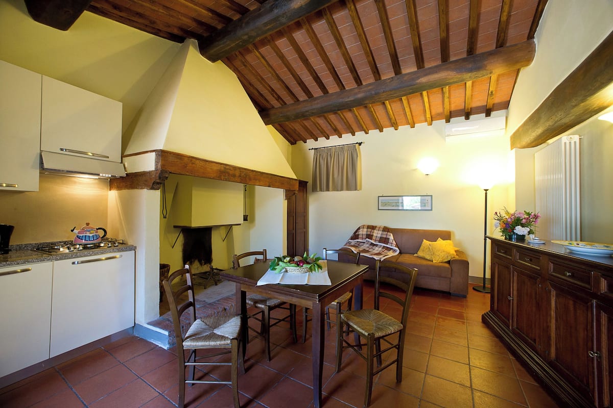 A compact kitchen with a dining area, wooden ceiling beams, and a cozy living space with a sofa and a lamp.