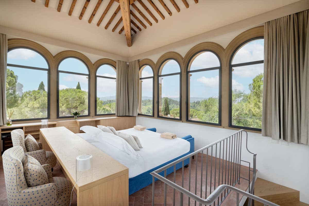 A bedroom with a large bed, a desk, and a chair. There are multiple windows with curtains, and a view of trees and mountains outside.