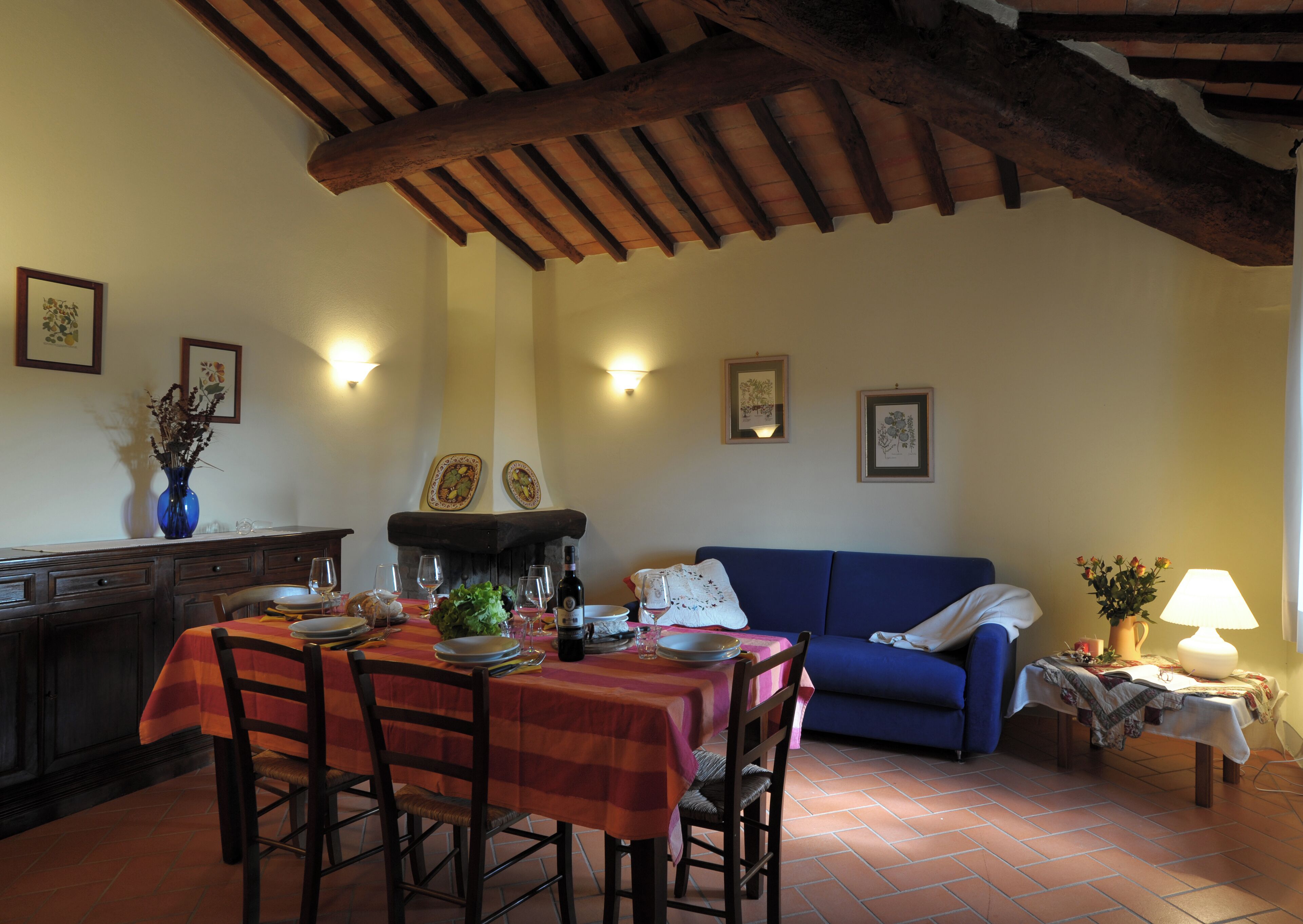 A dining area with a table set for four, a fireplace, and a blue sofa in a room with exposed wooden beams and tiled flooring.